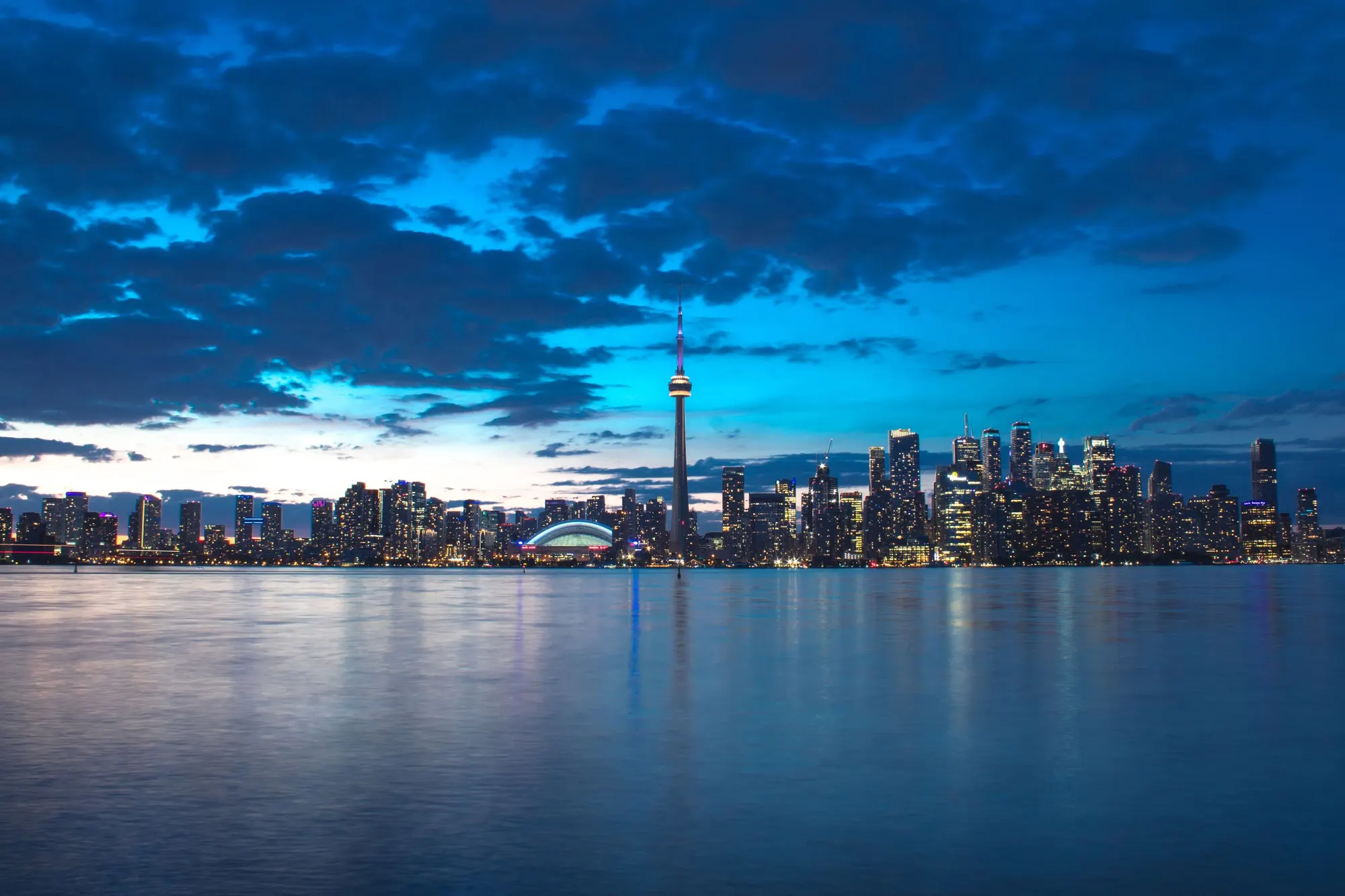 The Toronto skyline at blue hour seen across the water, with the CN Tower lit against heavy cloud